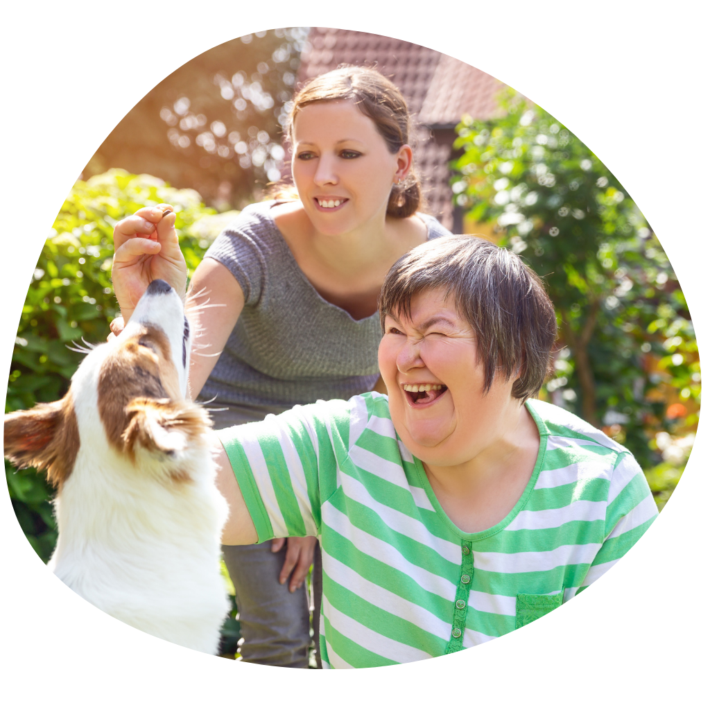 A young lady with learning disability accompanied by her female carer in the garden, laughing and playing with her dog.