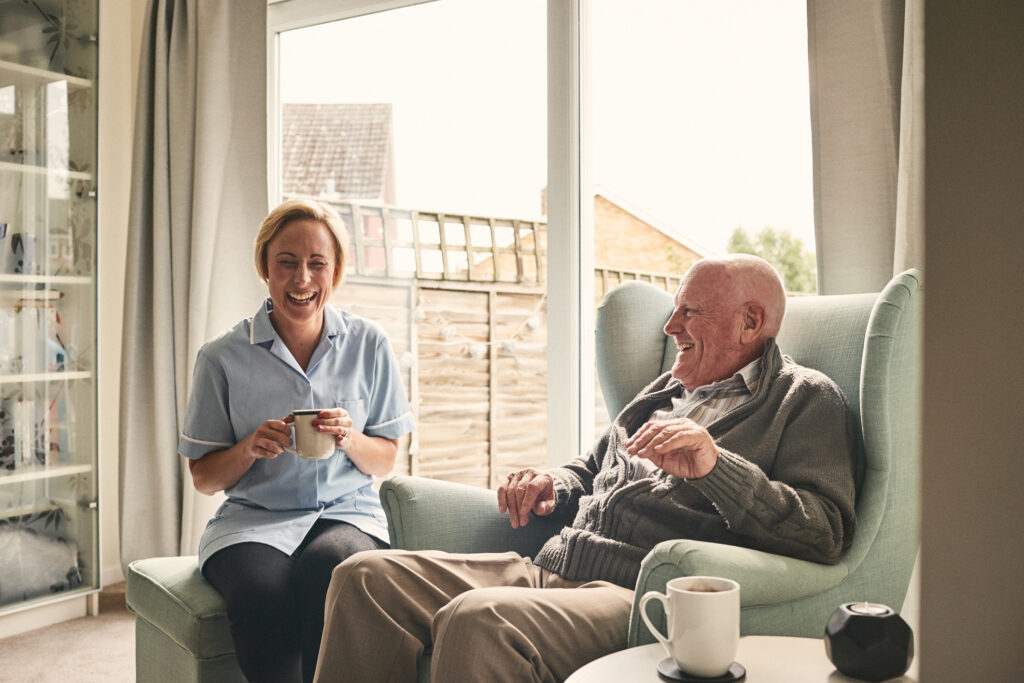 A gentleman in his armchair laughing with his carer, sat next to him.