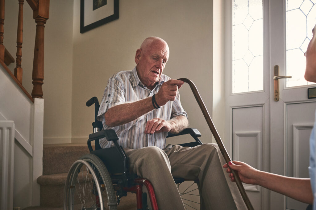 A gentleman in a wheelchair being assisted with his walking stick.